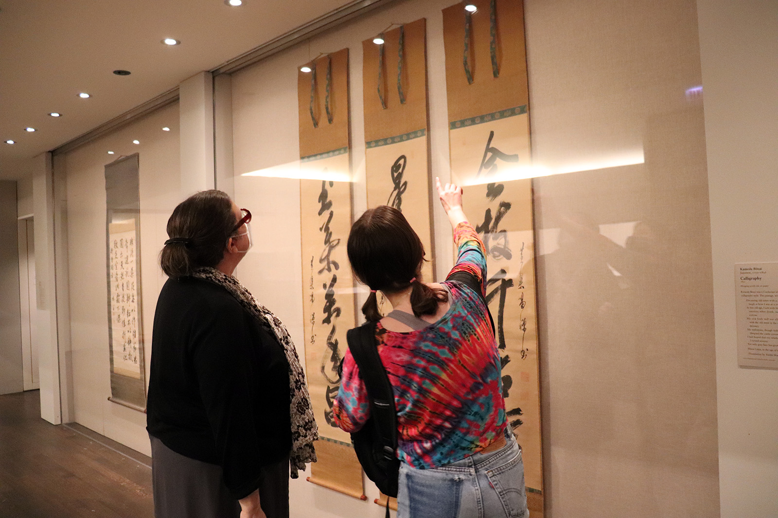 two women stand together poining and looking at three hanging banners displaying calligraphy by Japanese artist, Kameda Bōsai